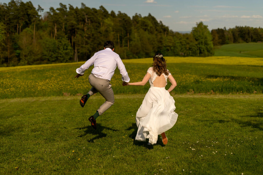 Ein Brautpaar steht Arm in Arm im Seepark Freiburg, lachend und glücklich an ihrem Hochzeitstag. Die Braut trägt ein elegantes Spitzenkleid und hält einen bunten Strauß, der Bräutigam einen dunkelblauen Anzug mit Fliege. Vor ihnen ein 'Just Married'-Banner. Ein Moment voller Freude, Leichtigkeit und echter Herzensmomente — eingefangen von Gregor Marstaller, Hochzeitsfotograf aus Freiburg im Breisgau.