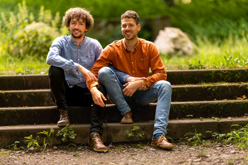 Zwei verlobte Männer lächeln authentisch bei einem Paarfotoshooting auf Steintreppe in Freiburg.