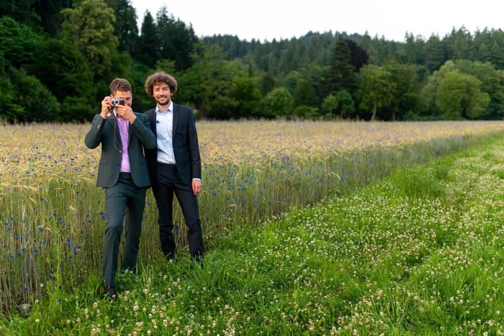 Zwei Männer in Anzügen bei Verlobungsshooting auf Wildblumenwiese vor Waldkulisse in Freiburg.