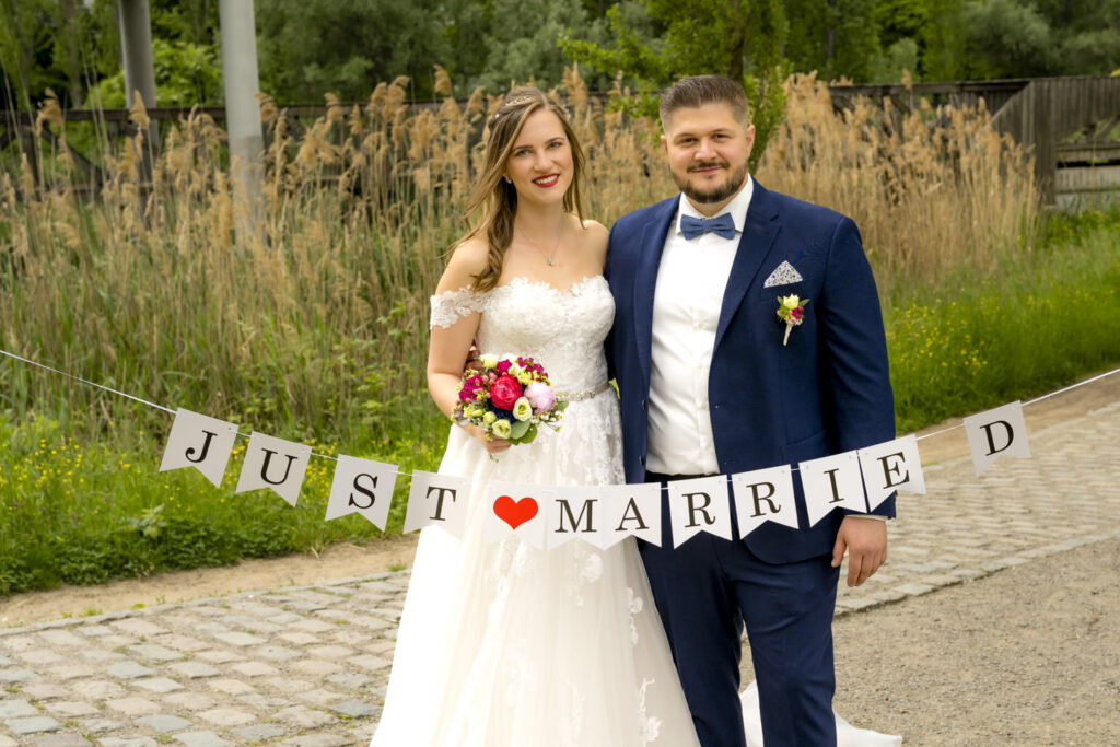 Hochzeitsfoto im Seepark Freiburg: Ein Brautpaar steht Arm in Arm auf einem Weg vor hohen Gräsern. Die Braut in einem weißen Spitzenkleid mit Blumenstrauß, der Bräutigam in dunkelblauem Anzug mit Fliege. Vor ihnen ein Banner mit der Aufschrift 'Just Married'. Fotografiert von Gregor Marstaller.
