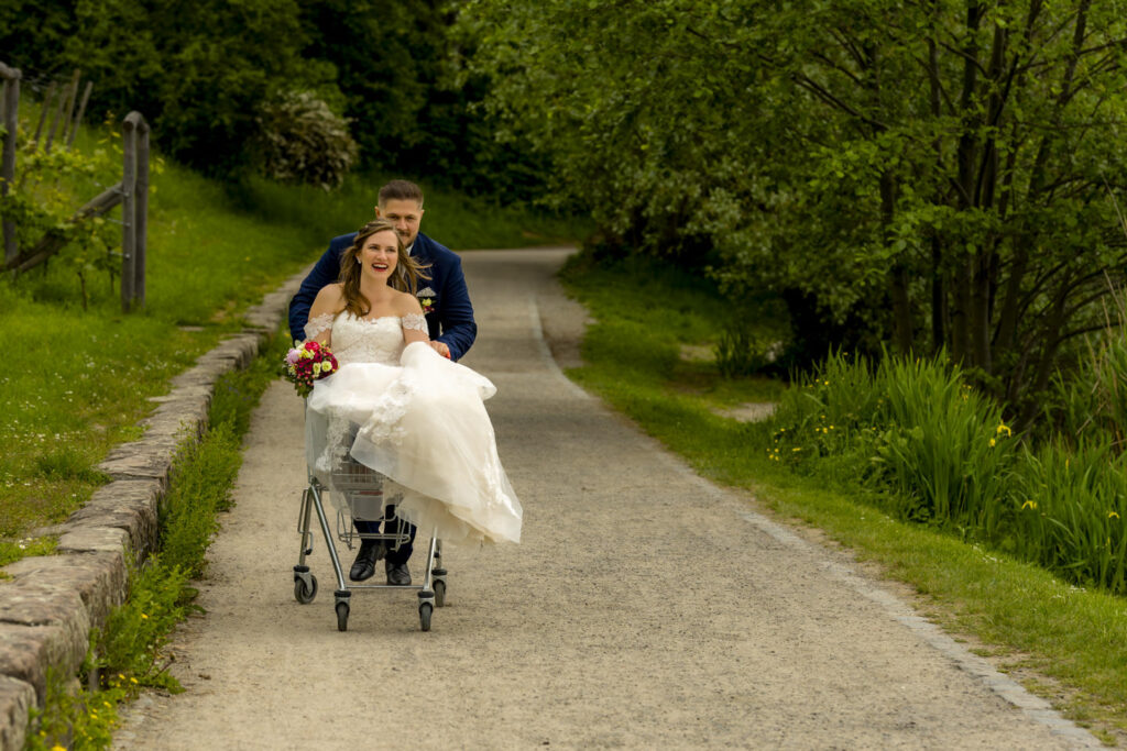 Braut in weißem Hochzeitskleid sitzt lachend in einem Einkaufswagen, während der Bräutigam sie auf einem Waldweg schiebt. Beide genießen den Moment voller Freude und Leichtigkeit.