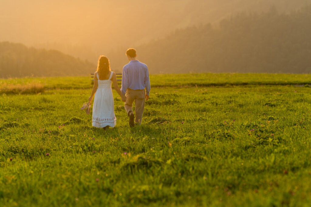 Brautpaar Hand in Hand auf weitem Feld bei goldenem Sonnenuntergang mit nebligen Hügeln.