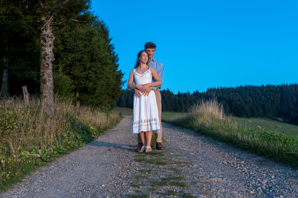 Brautpaar umarmt sich auf Landstraße bei goldenem Sonnenuntergang in Berglandschaft.