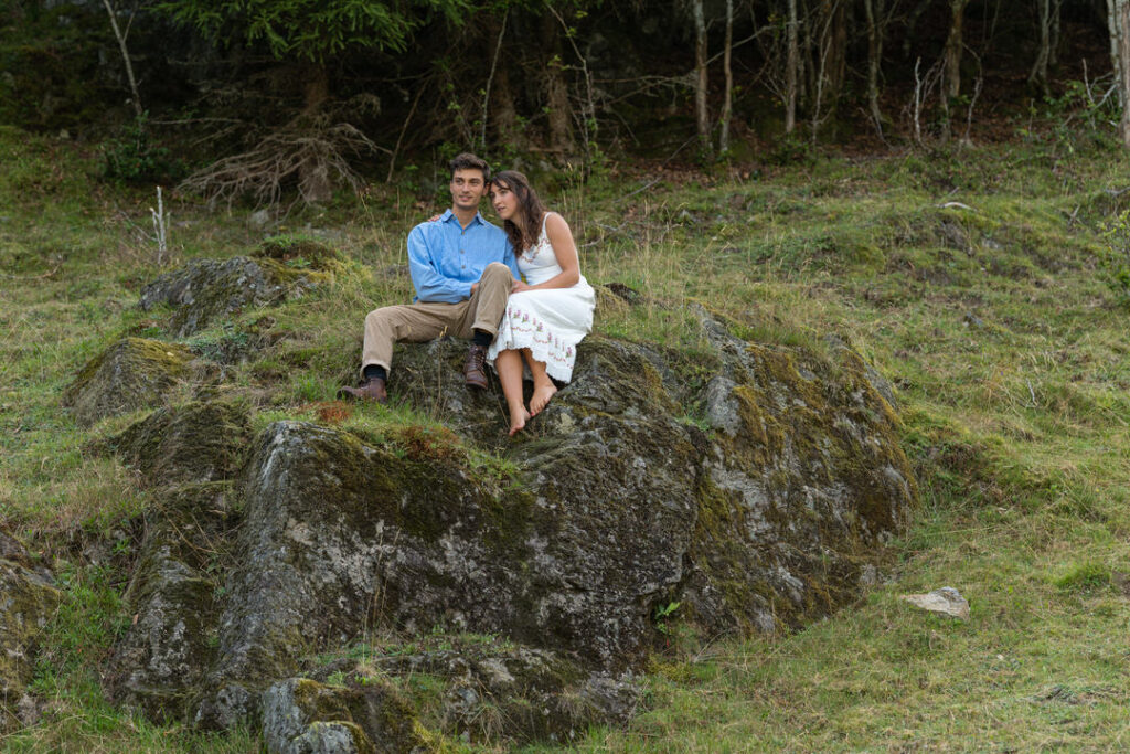 Verliebtes Paar sitzt auf moosbedecktem Felsen im nebligen Wald während romantischem Fotoshooting.