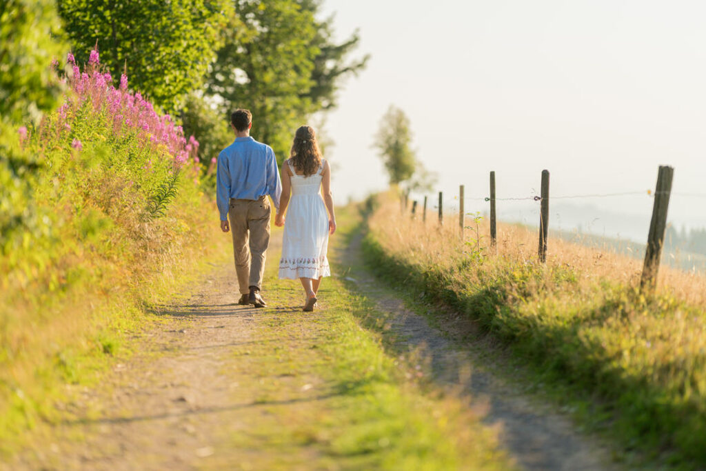 Brautpaar spaziert Hand in Hand auf ländlichem Feldweg im goldenen Sonnenlicht des Sonnenuntergangs.