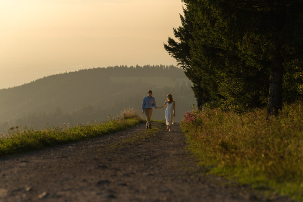 Paar wandert mit weißem Hund durch goldene Abendlandschaft auf Naturpfad in den Bergen.
