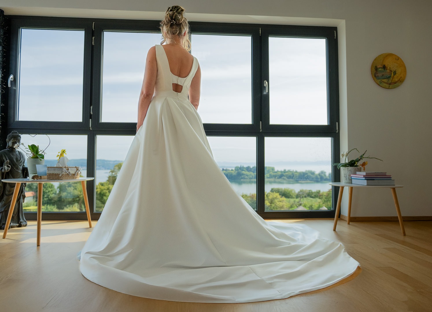 Braut in weißem Hochzeitskleid mit Schleppe vor Panoramafenster mit Bodensee-Blick beim Getting Ready.