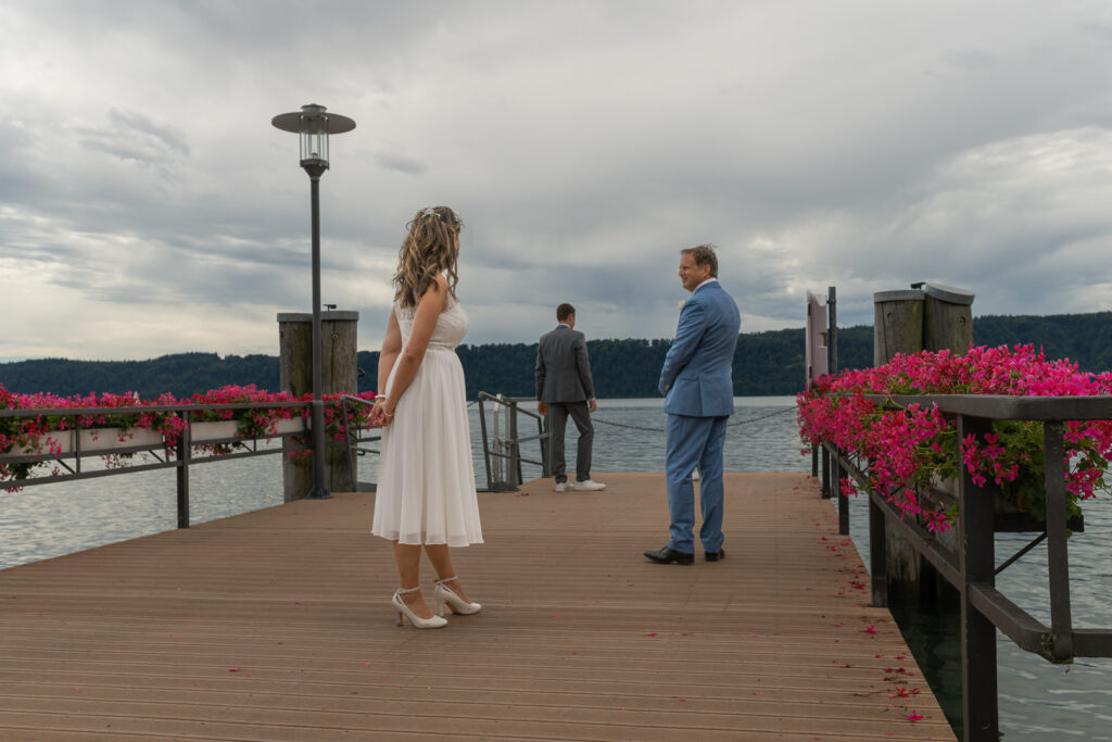 Brautpaar auf Holzsteg am Bodensee mit pinken Blumen bei bewölktem Himmel während Hochzeitsreportage.