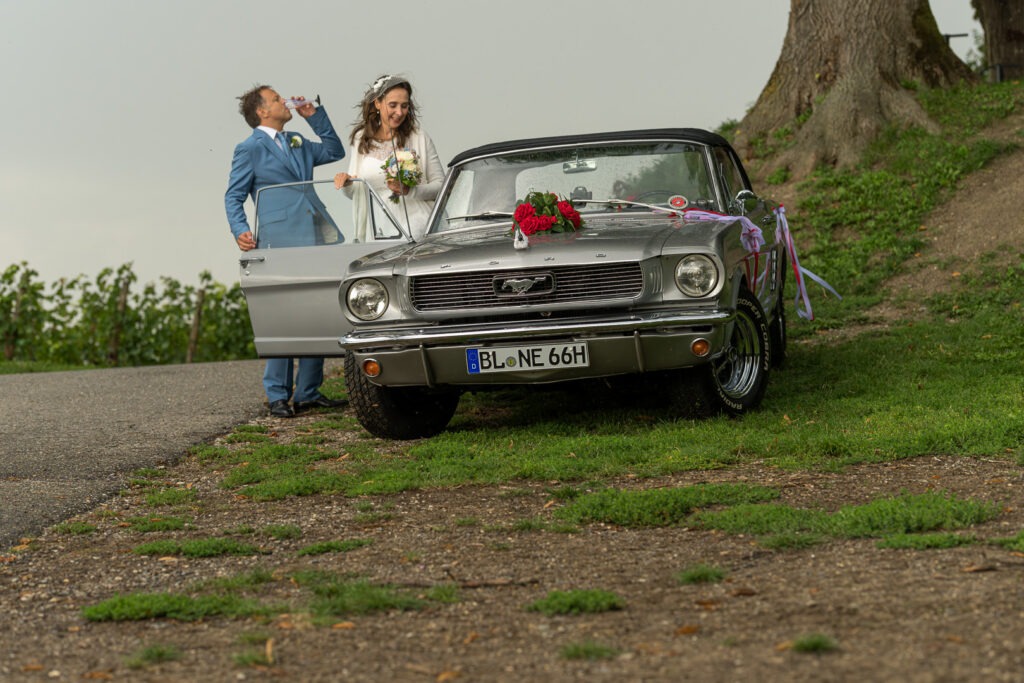 Brautpaar mit klassischem Ford Mustang Cabrio als Hochzeitsauto auf ländlichem Weg mit roter Schleife.