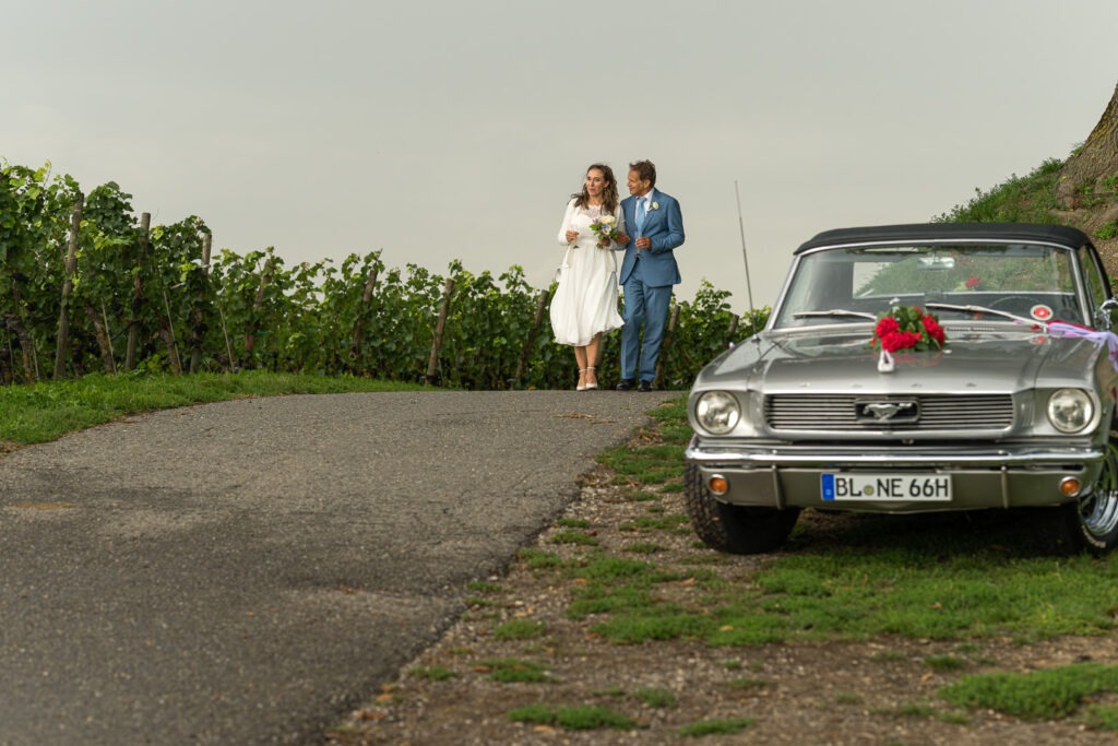 Brautpaar mit geschmücktem Ford Mustang Oldtimer als Hochzeitsauto im Weinberg bei romantischem Fotoshooting.