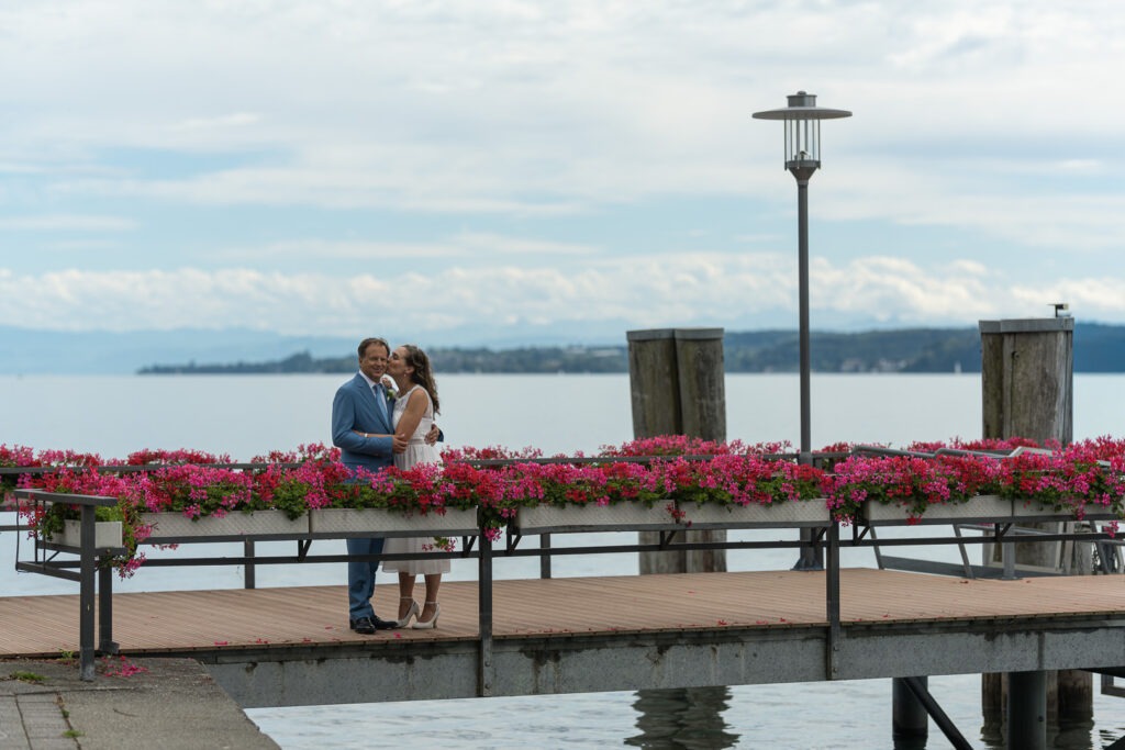 Brautpaar auf blumengeschmückter Seebrücke am Bodensee mit Bergpanorama im Hintergrund.