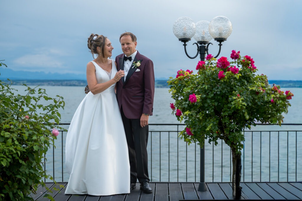 Brautpaar in eleganter Hochzeitskleidung auf Seeterrasse bei Sonnenuntergang mit dramatischem Himmel und Blumendekoration.