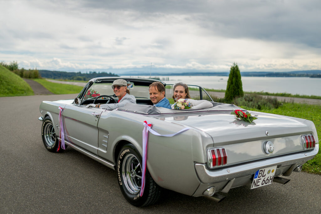 Silbernes Ford Mustang Cabrio als Hochzeitsauto am Seeufer mit Brautpaar und rosa Dekoration.