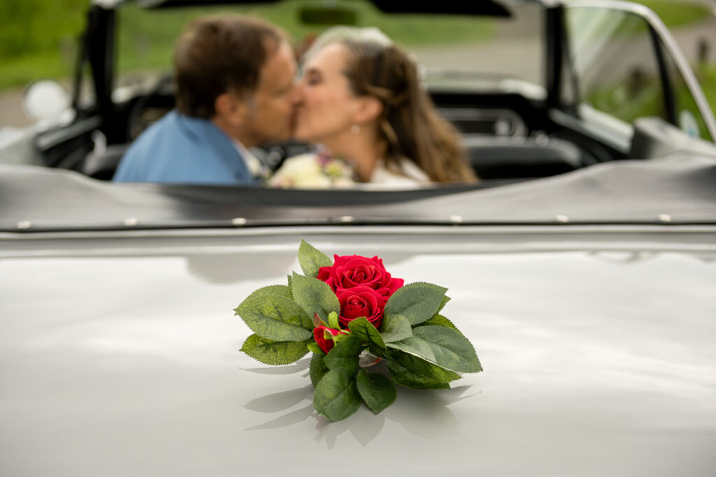 Brautpaar küsst sich im weißen Hochzeits-Cabrio, dekoriert mit roter Rose auf der Motorhaube.