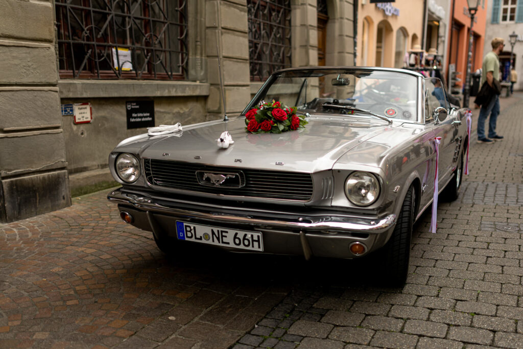 Silberner Ford Mustang Oldtimer mit roter Schleife als Hochzeitsauto auf historischem Kopfsteinpflaster in deutscher Altstadt.