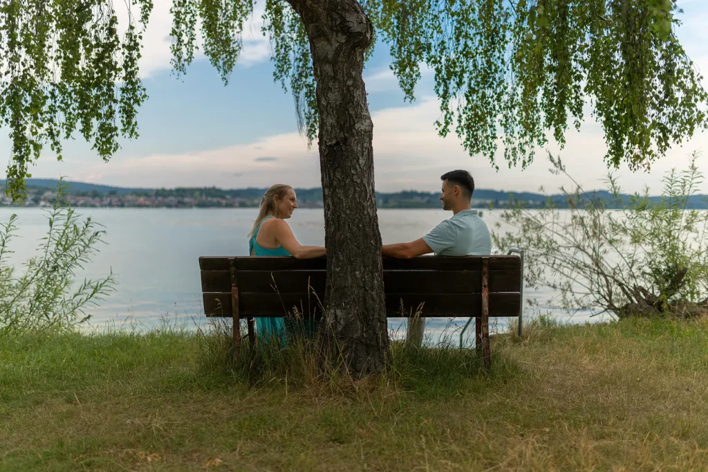 Paar Svenja und Yannik sitzen auf Bank am Bodensee unter Weide.